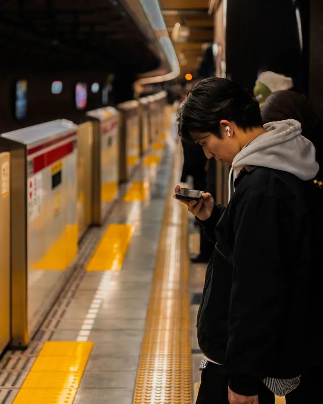 a commuter bows to his phone while the platform screen doors recede into dark