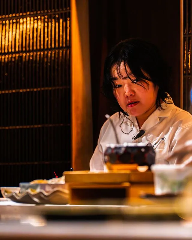the chef at the counter in warm amber light, a mic clipped to her collar